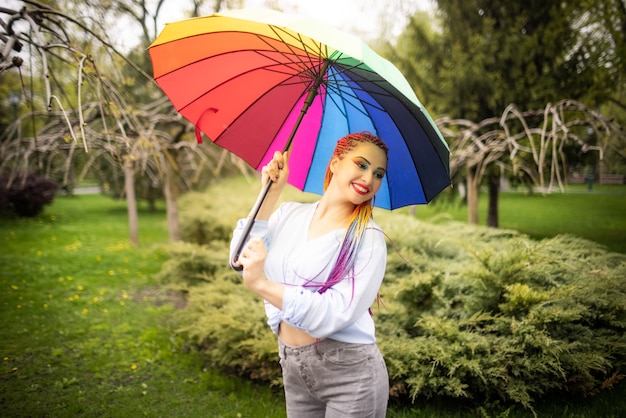 Girl in a bluish shirt with bright makeup and long colored braids. Smiling and holding an umbrella in the colors of the rainbow on the background of a flowered park enjoying the coming spring.