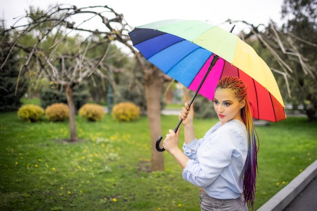 Девушка в голубоватой рубашке с ярким макияжем и длинными разноцветными косами. Улыбается и держит зонтик