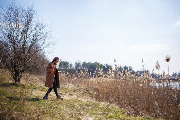 A girl in a brown coat, hat and glasses walks in a park with a lake under the bright sun. Drinking tea from a paper cup. The beginning of spring