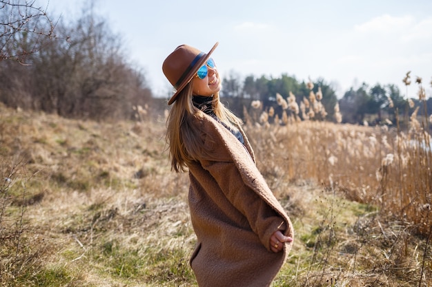 A girl in a brown coat, hat and glasses walks in a park with a lake under the bright sun. Rejoices in life and smiles. The beginning of spring