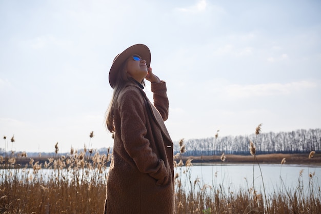 A girl in a brown coat, hat and glasses walks in a park with a lake under the bright sun. Rejoices in life and smiles. The beginning of spring