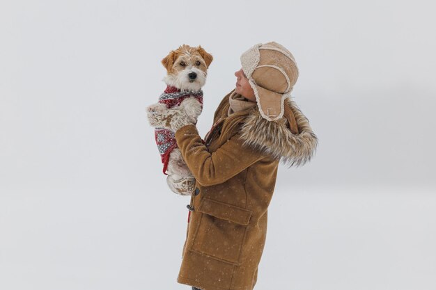 A girl in a brown coat and a jack russell terrier stand on a white background during a blizzard The dog sits on the hands of a man Christmas concept