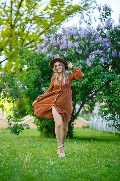 A girl in a brown hat and dress walks through the park, she has a European appearance and a smile on her face.