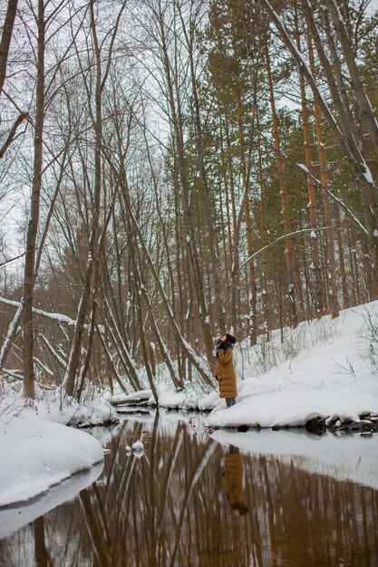 A girl in a brown jacket stands near the river of the winter forest looking up. Winter forest. Winter river.