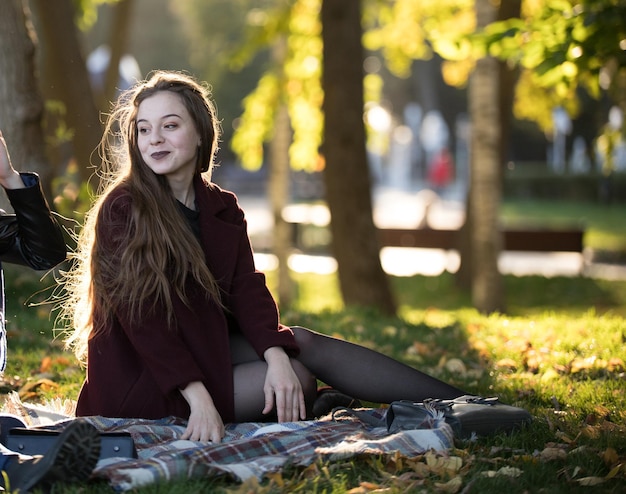 Girl in a burgundy coat sitting on a plaid in the autumn sunny park