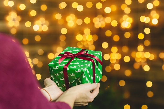 A girl in a burgundy sweater, holds in her hands a green box gift with a red ribbon. In the background is a wooden wall with a garland of stars. Concept on the theme of holidays.