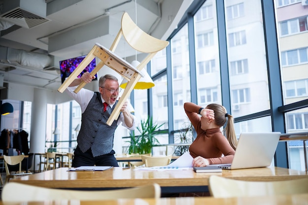 Girl business lady sitting at a wooden table with a laptop and working teacher boss mentor indicates her mistakes. Screams and hits the table with his hand