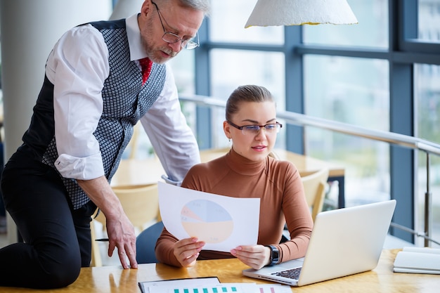 Girl business woman sitting at a wooden table with a laptop and discussing a new project with her mentor boss teacher. The man is fooling around. New business development concept