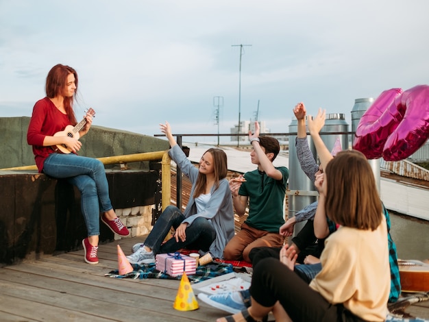 Girl busker playing ukulele at a rooftop. Artistic musician lifestyle. Teenagers leisure. Friends enjoy music with hands in the air