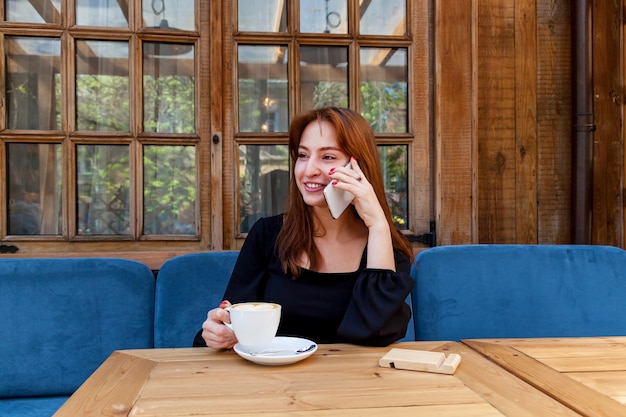 Photo a girl in a cafe at a table talking on the phone girl in a black dress talking on the phone over a cup of coffee