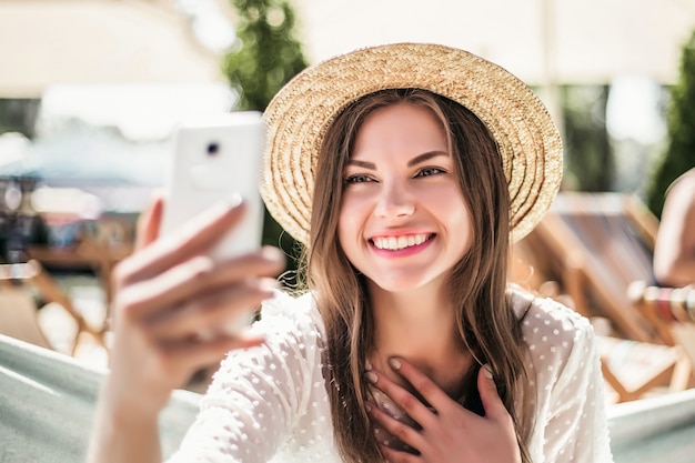 Girl calling by videoconference while sitting on the beach in summer in a straw hat is embarrassed and smiling