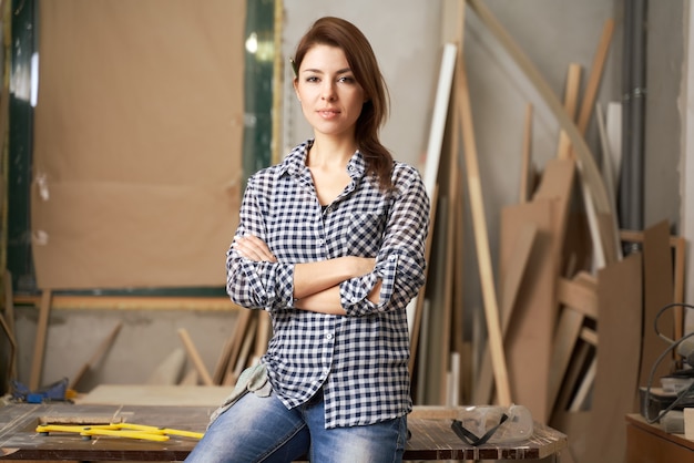 Girl carpenter in checkered shirt with arms crossed in workshop