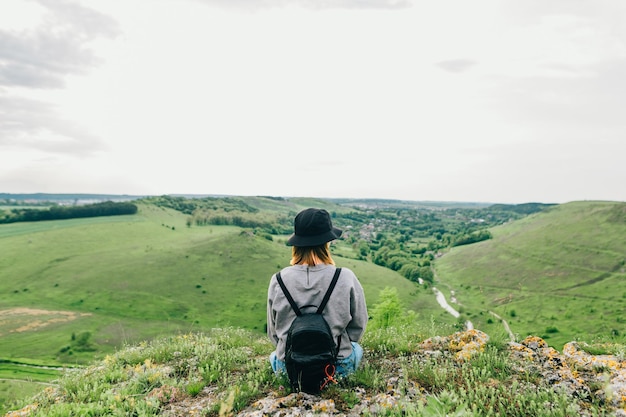 カジュアルな服を着た女の子が丘の上の岩の上に座って、美しいウクライナの風景を見ています
