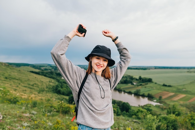 girl in casual clothes stands on a hill with a smartphone in hand and rejoices with raised hands