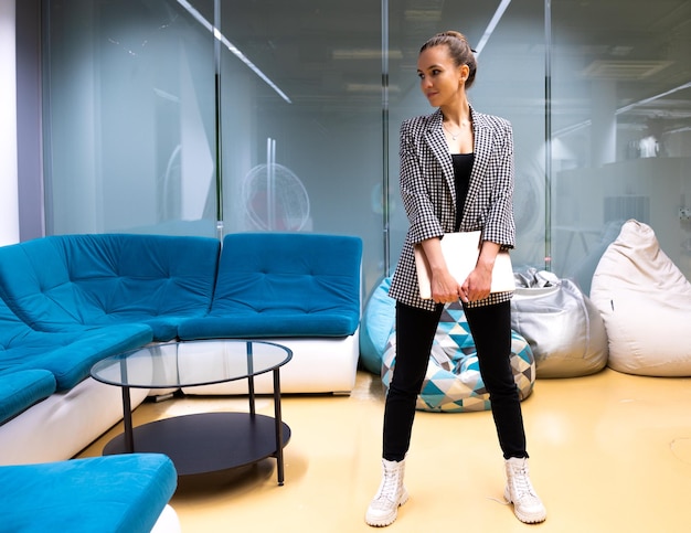 A girl in casual style stands with a laptop in the office space. Looks away and smiles.