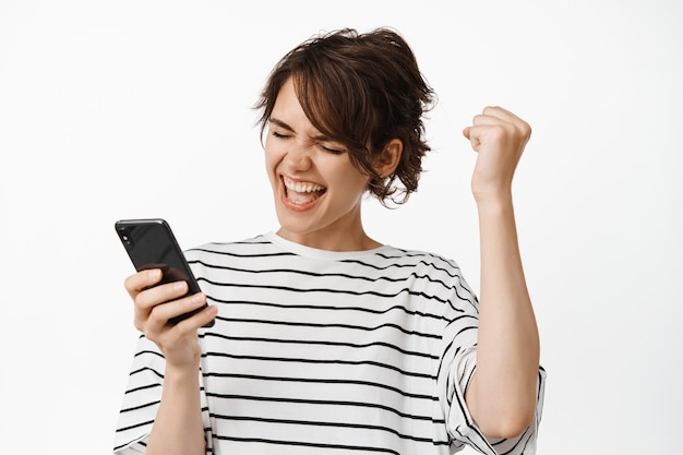 Girl celebrating, looking at mobile phone and rejoicing, scream from happy achievement on smartphone app, standing on white