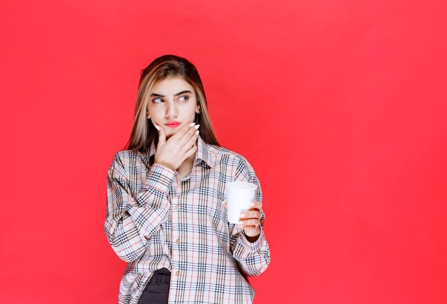 Girl in checked shirt holding a white disposable coffee cup and looks thoughtful or has good ideas