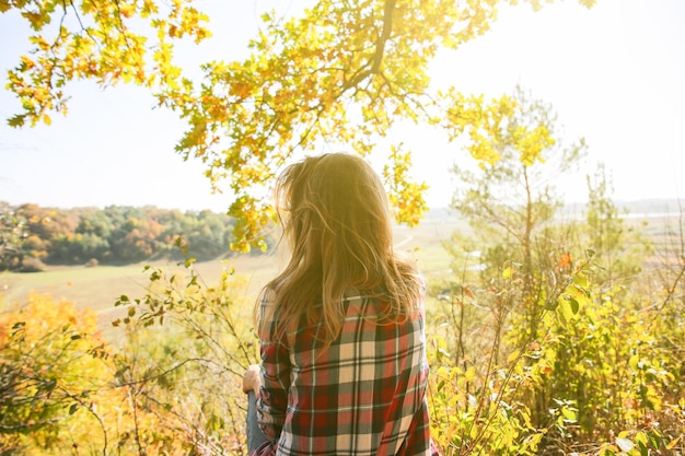 チェッカーシャツを着た女の子が秋の森に座っている季節のコンセプトスタイリッシュな流行に敏感な服を屋外で黄色の葉や木の近くの美しい女性の周りの自然哲学