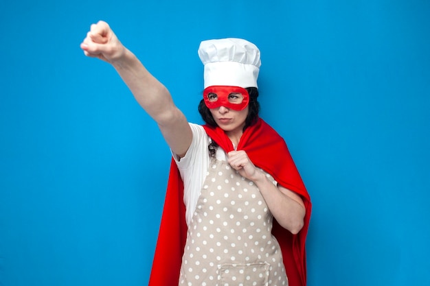 Girl chef in superman costume holds kitchen item on blue background woman housewife