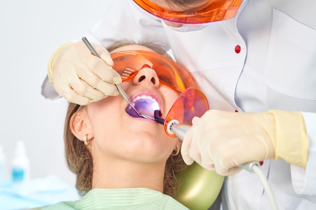 Девочки у врача. Стоматолог помещает пломбу на зуб с помощью зубной полимеризационной лампы в полости рта. на фоне клиники