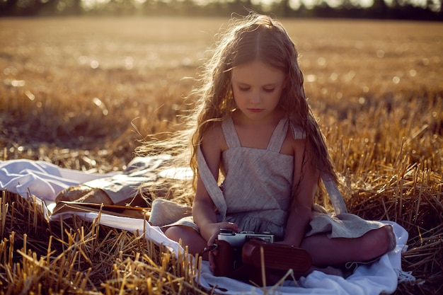 Girl child in a dress sitting on a mown field with a camera on a blanket with bread and a book