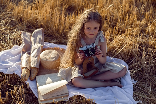 Girl child in a dress sitting on a mown field with a camera on a blanket with bread and a book