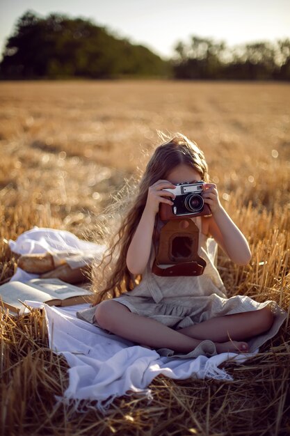 Photo girl child in a dress sitting on a mown field with a camera on a blanket with bread and a book