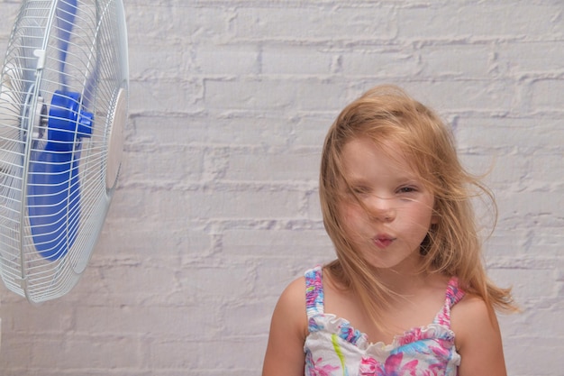 The girl child in front of an electric fan enjoys the flow of cool wind on a hot summer day