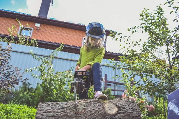 The girl child in a helmet with an electric saw saws a log