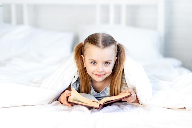A girl child is reading a book on the bed at home on a white cotton bed under a blanket and smiles sweetly