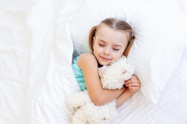 Девочка спит на кровати дома на белой хлопчатобумажной кровати, обнимая в руках игрушку плюшевого мишку и сладко улыбаясь во сне.