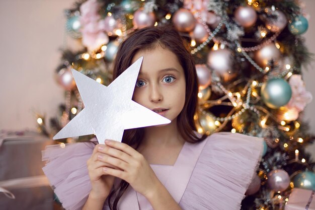 Girl child in a pink dress sits at the Christmas tree and holds a star in her hands near her face