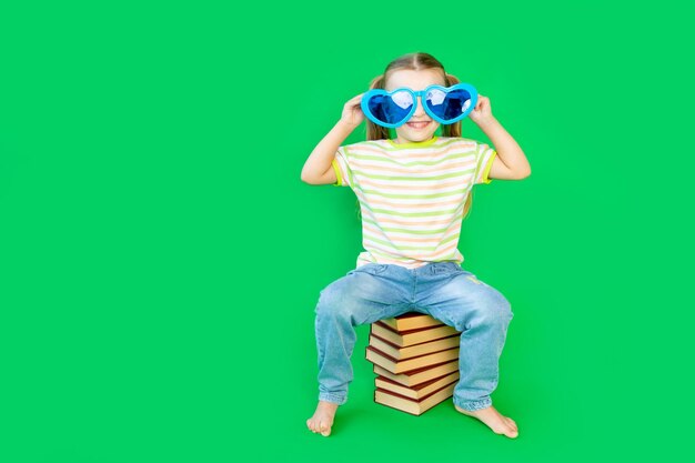 A girl child sits on a green background on books in blue big funny glasses and smiles space for text