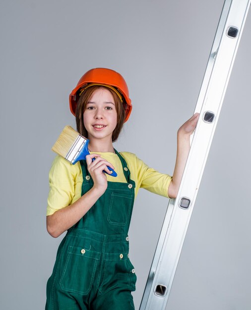 Girl child wearing uniform and helmet while working in workshop with painting brush on ladder labor day