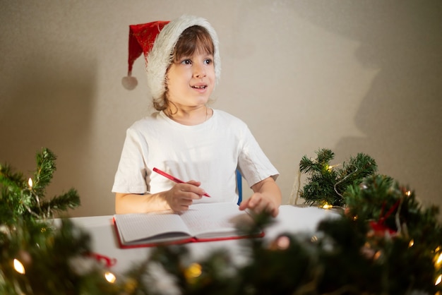 Photo girl child in a white t-shirt with a red pen and a new year's cap on his head writes dear santa a letter at the table in anticipation of the birth. wish list of gifts for the new year.