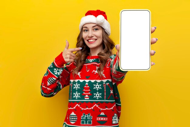 girl in christmas sweater and santa claus hat shows blank smartphone screen on yellow background