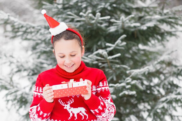 Girl in Christmas sweater and Santa hat rim with gift in winter forest