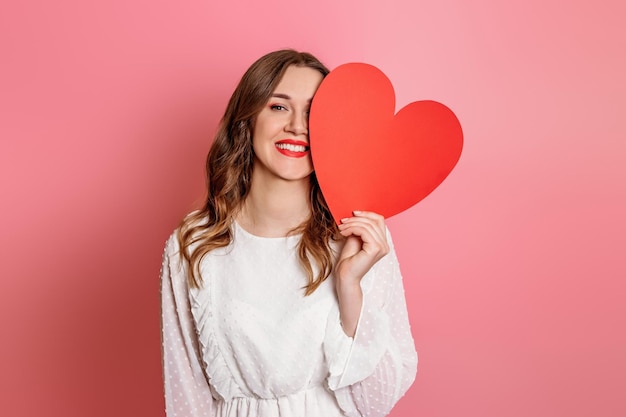 Girl closes one eye with a big paper heart isolated on a pink background Valentine's Day