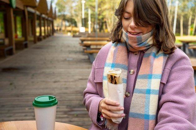 コートを着た女の子が屋外カフェのテーブルに立ち、屋台の食べ物を食べる