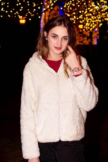 A girl in a coat and gloves stands under a Christmas tree decorated with lights. New Year 2022.