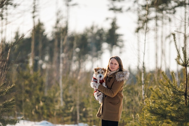 コートを着た女の子が、ジャック・ラッセル・テリア犬を両腕に抱え、雪のクリスマス・コンセプトの冬のトウヒの森で