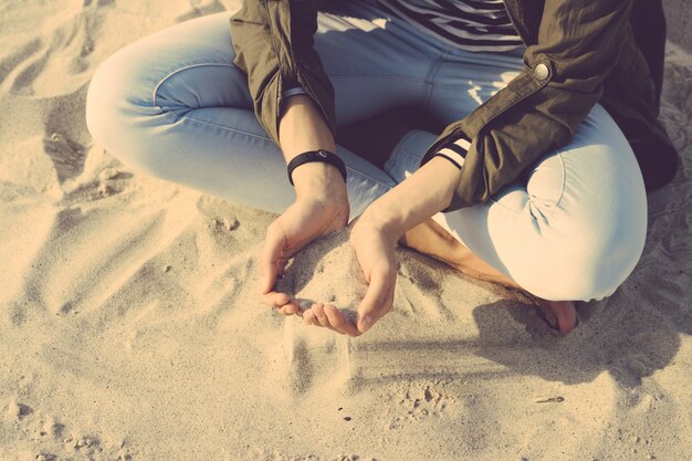 Girl in coat and jeans sitting on the beach cross-legged, in her palms sand