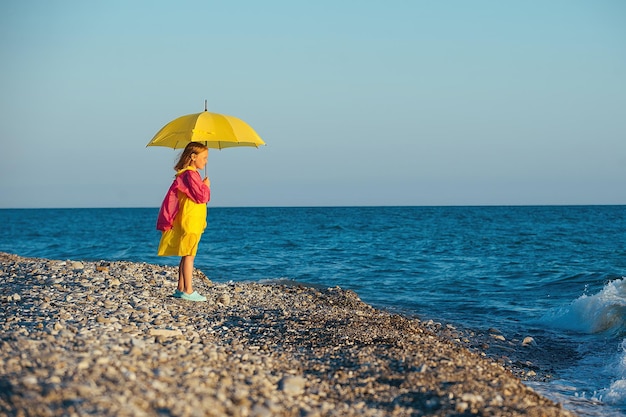 A girl in a colored raincoat and with a yellow umbrella stands
on the shore of the blue sea