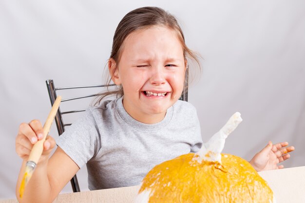 a girl cries and makes a handmade papier-mache pumpkin for Halloween on a gray wall,