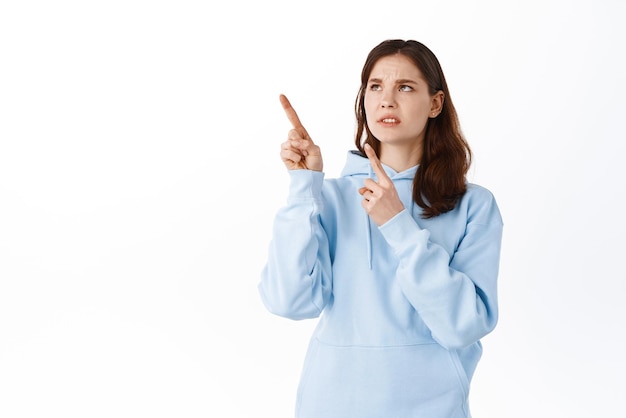 Girl cringe from something disgusting Displeased young woman pointing and looking at upper left corner with aversion and dislike grimacing standing over white background