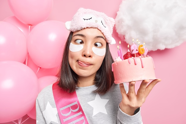 Foto ragazza incrocia gli occhi fa una smorfia divertente lecca le labbra applica cerotti al collagene per ridurre le rughe festeggia il compleanno da sola tiene una torta dolce