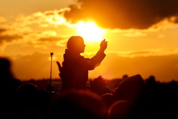 Ragazza sopra la folla contro il cielo al tramonto. divertendosi e utilizzando il suo smartphone al festival estivo.