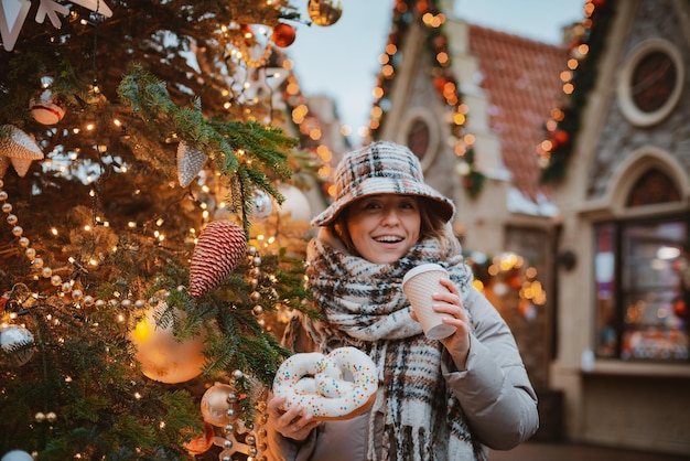 Ragazza tazza bevanda calda mentre si cammina nel mercatino di natale decorato con luci natalizie la sera. felice nella grande città.