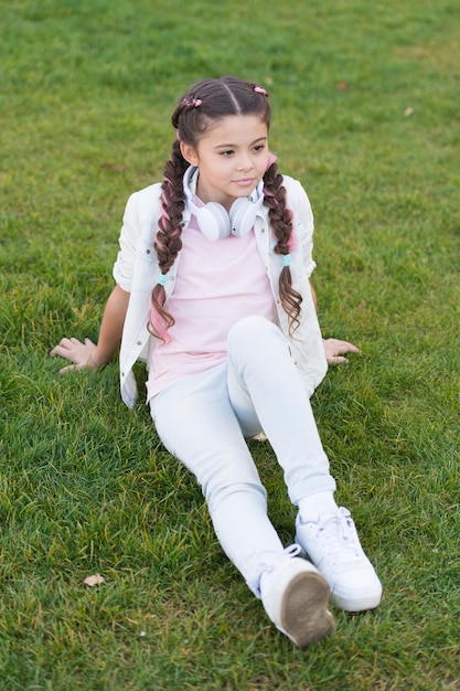 Girl cute kid green grass background Healthy emotional happy kid relaxing outdoors What makes child happy Girl braids hairstyle and modern headphones enjoy relax Secrets to raising happy child