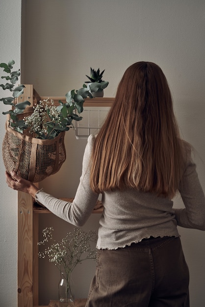 The girl decorates a wooden rack with an eco-bag with eucalyptus branches, flowers, houseplants